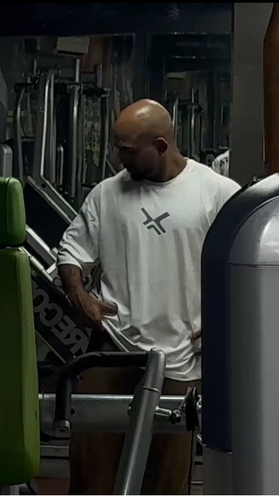 A man in a white t-shirt exercises on a gym machine inside a gym with green and black weight machines.